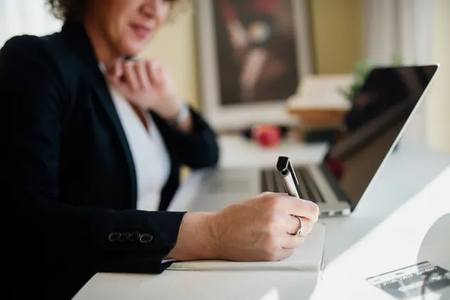 A business professional is seated at a sleek desk, actively taking notes with a pen while focused on a laptop screen. Natural light enhances the workspace's ambiance. Business email pricing: plans, fees and buying questions
